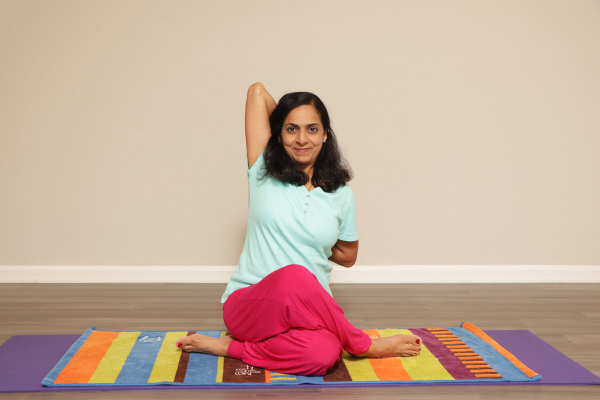 A woman performs yoga on a colorful mat in a room with a beige wall. She is sitting on the floor with her left arm raised behind her head and her right hand resting on her back. A person wearing red pants is lying flat on the mat in front of her, with their head turned to the side. The woman has black hair, a light blue shirt, and a smile.